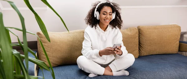 Panoramic orientation of happy african american girl in wireless headphones using smartphone while sitting on sofa — Stock Photo