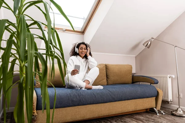 Selective focus of positive african american girl in wireless headphones using smartphone while sitting on sofa — Stock Photo