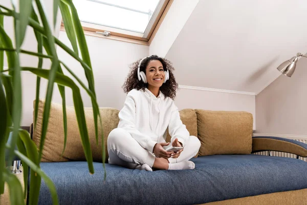 Selective focus of happy african american girl in wireless headphones holding smartphone while sitting on sofa — Stock Photo