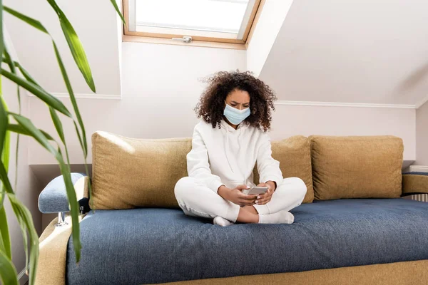 Enfoque selectivo de rizado chica afroamericana en máscara médica usando teléfono inteligente en la sala de estar - foto de stock