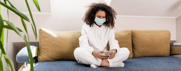 Imagen horizontal de la chica americana africana rizada en máscara médica usando teléfono inteligente en la sala de estar - foto de stock