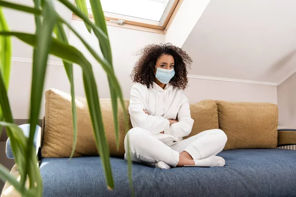 Selective focus of curly african american girl in medical mask sitting with crossed arms on sofa — Stock Photo