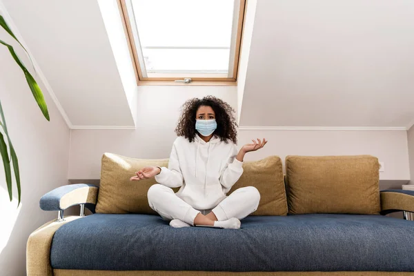 Selective focus of curly african american girl in medical mask gesturing while sitting on sofa — Stock Photo