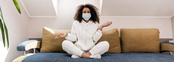 Horizontales Konzept eines lockigen afrikanisch-amerikanischen Mädchens mit medizinischer Maske, das auf dem Sofa gestikuliert — Stockfoto