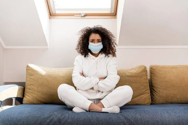 Кудрявая африканская американка в медицинской маске сидит со скрещенными руками на диване — стоковое фото