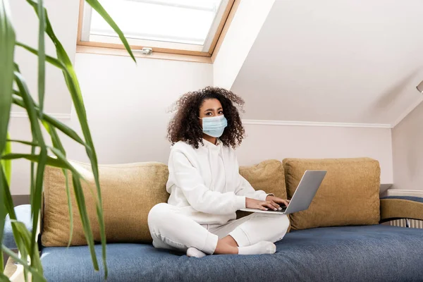 Selective focus of curly african american freelancer in medical mask using laptop — Stock Photo
