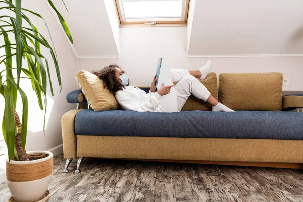 Curly african american woman in medical mask lying on sofa and reading magazine — Stock Photo