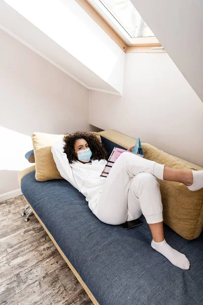 Mulher afro-americana em máscara médica deitada no sofá e revista de leitura — Fotografia de Stock