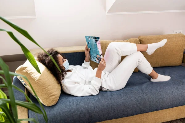 Foyer sélectif de la femme afro-américaine bouclée dans le masque médical couché sur le canapé et la lecture magazine — Photo de stock
