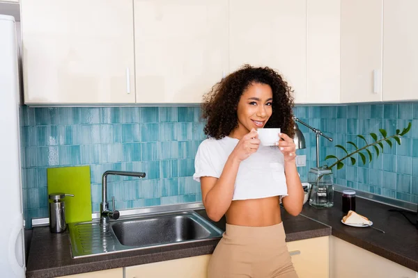 Felice ragazza afro-americana guardando la fotocamera e tenendo in mano una tazza di caffè — Foto stock