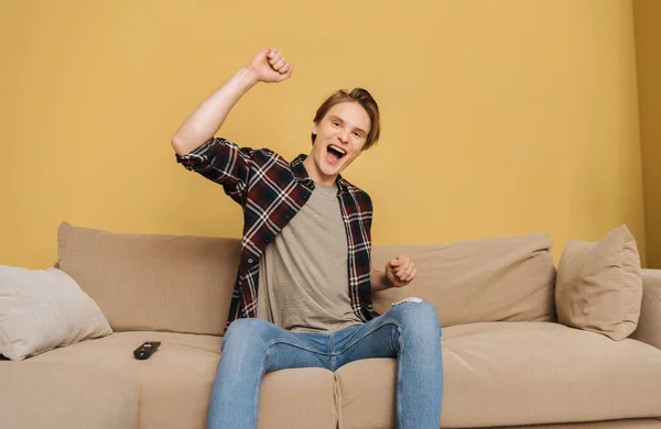 Hombre excitado con la boca abierta sentado en el sofá cerca del mando a distancia, fin del concepto de cuarentena - foto de stock