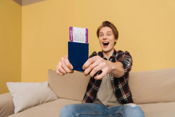 Selective focus of happy man with opened mouth sitting on sofa and holding passport with air ticket, end of quarantine concept — Stock Photo