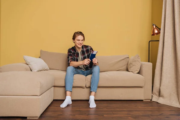 Fröhlicher Mann sitzt auf Sofa und hält Reisepass mit Flugticket in der Hand, Ende der Quarantäne — Stockfoto