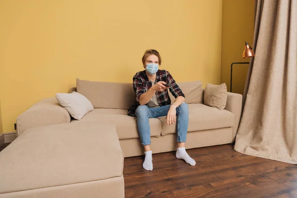 Man in medical mask sitting on sofa and holding remote controller — Stock Photo