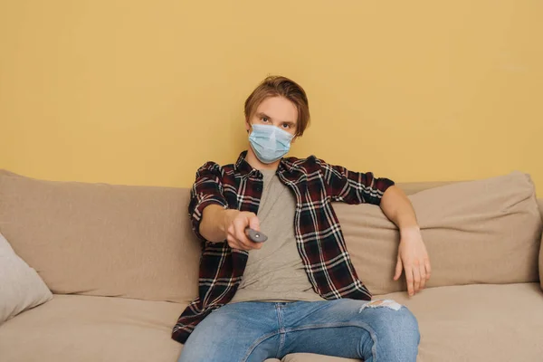 Junger Mann mit medizinischer Maske sitzt auf Sofa und hält Fernbedienung in der Hand — Stockfoto