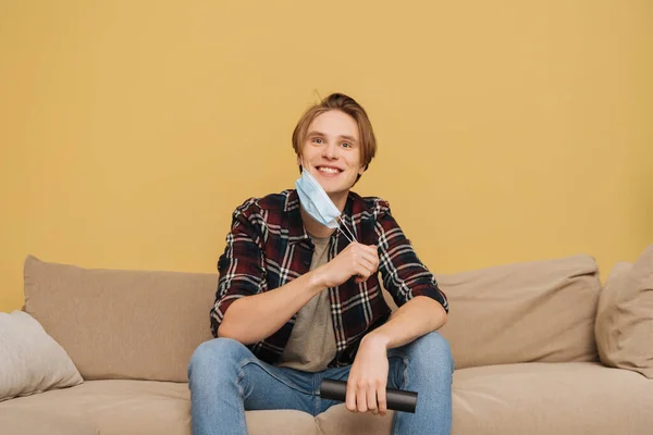 Glücklicher Mann, der medizinische Maske berührt, auf Sofa sitzt und Fernbedienung hält, Ende des Quarantäne-Konzepts — Stockfoto