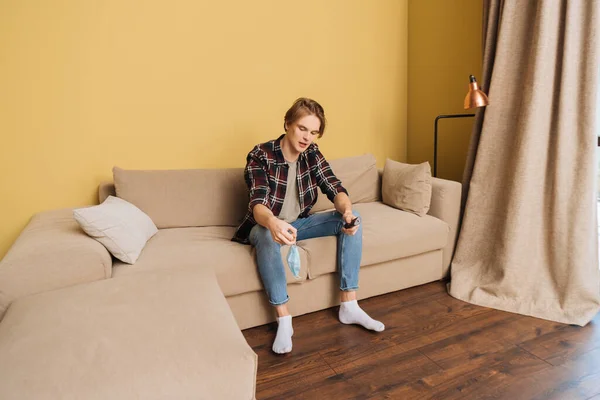 Hombre sentado en el sofá, la celebración de la máscara médica y el mando a distancia, fin del concepto de cuarentena - foto de stock