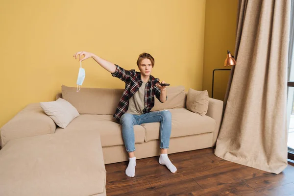 Young man sitting on sofa, holding medical mask and remote controller, end of quarantine concept — Stock Photo