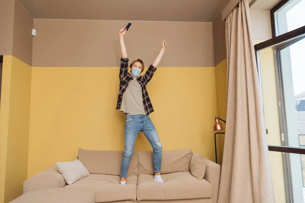 Man in medical mask standing on sofa and holding remote controller above head, end of quarantine concept — Stock Photo