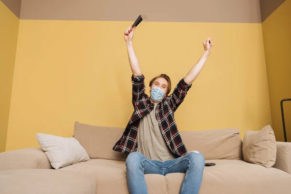 Hombre en máscara médica sentado en el sofá y la celebración de mando a distancia por encima de la cabeza, el final del concepto de cuarentena - foto de stock