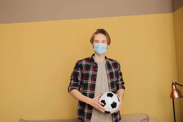 Junger Mann mit medizinischer Maske hält Fußball in der Hand und blickt in die Kamera — Stockfoto
