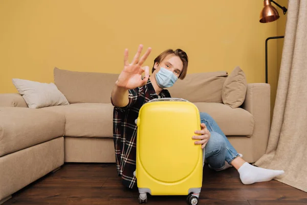 Вибірковий фокус чоловіка в медичній масці, що сидить на підлозі біля багажу і показує знак ок, кінець концепції карантину — стокове фото