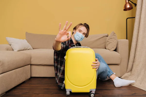 Selektiver Fokus des Mannes mit medizinischer Maske, der auf dem Boden in der Nähe des Gepäcks sitzt und OK-Zeichen zeigt, Ende des Quarantäne-Konzepts — Stockfoto