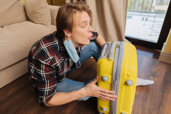 Young man in medical mask sitting on floor and blowing on dust on baggage, end of quarantine concept — Stock Photo