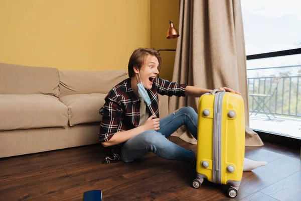 Excited man in medical mask sitting on floor and looking at baggage, end of quarantine concept — Stock Photo