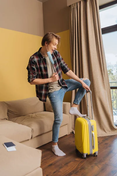 Homme heureux en masque médical debout près des bagages et passeport sur le canapé, fin du concept de quarantaine — Photo de stock