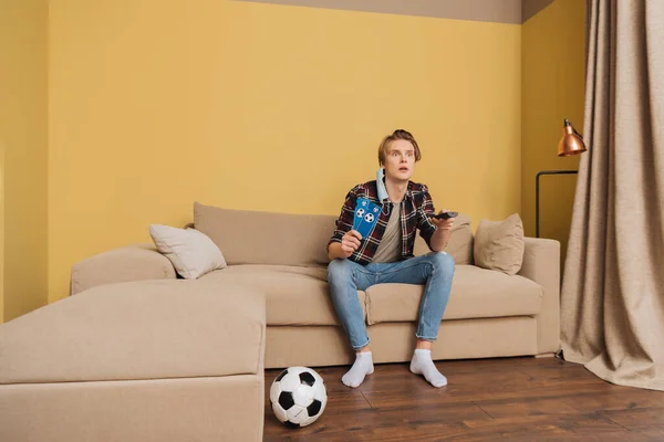 Homem emocional em máscara médica segurando bilhetes jogo esporte perto de futebol enquanto assiste campeonato em casa — Fotografia de Stock