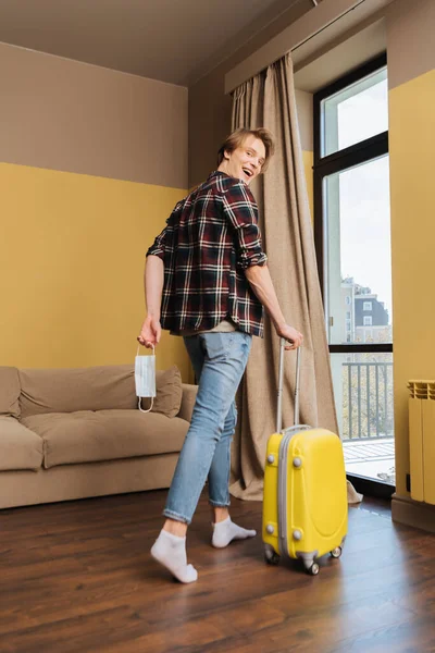 Счастливый человек, держащий медицинскую маску и идущий с багажом, конец карантинной концепции — стоковое фото