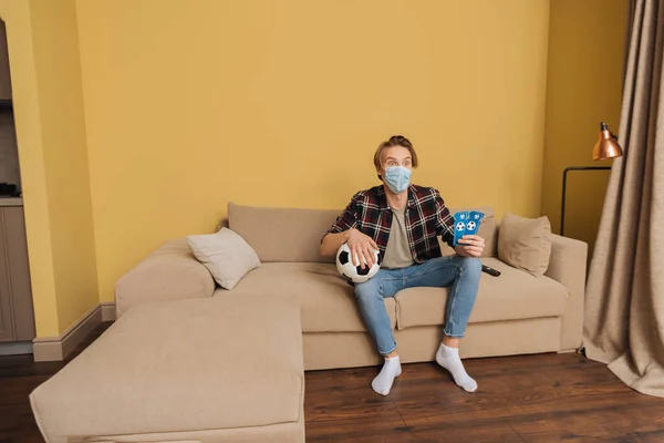 Überraschter Mann mit medizinischer Maske hält Fußball- und Sportspielkarten in der Hand, während er zu Hause die Meisterschaft verfolgt — Stockfoto