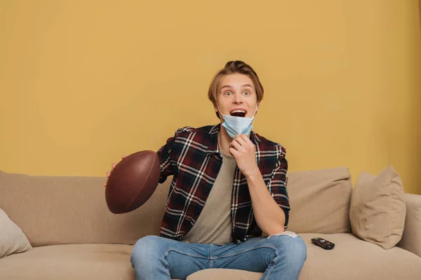 Excited young man in medical mask holding american football while watching championship in living room — Stock Photo