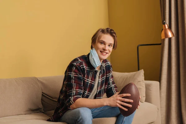 Felice giovane uomo in maschera medica che tiene il calcio americano in soggiorno — Foto stock