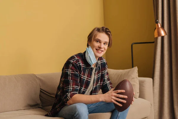 Happy young man in medical mask holding american football in living room — Stock Photo
