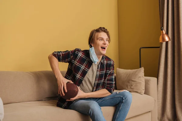 Excited man in medical mask holding american football while watching championship in living room — Stock Photo