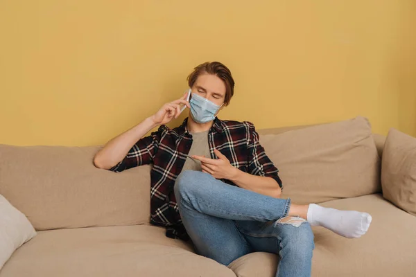 Homme en masque médical gestuelle tout en parlant sur smartphone dans le salon — Photo de stock