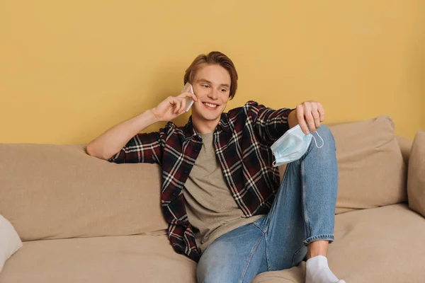 Handsome holding medical mask and smiling while talking on smartphone in living room — Stock Photo