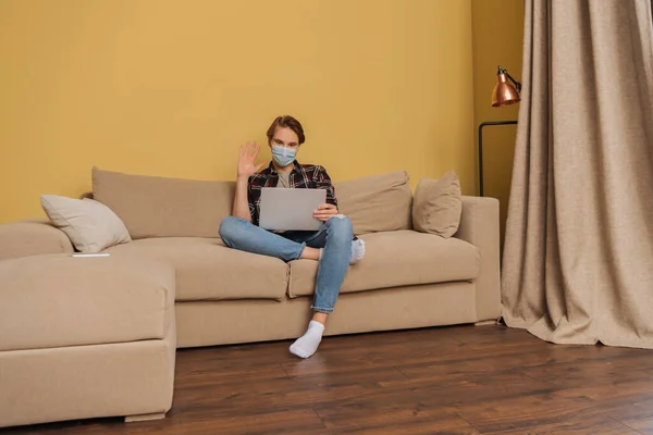 Homem em máscara médica acenando a mão enquanto tem vídeo chat na sala de estar — Fotografia de Stock