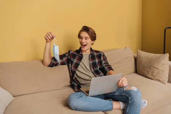 Happy freelancer holding medical mask near laptop in living room, end of quarantine concept — Stock Photo