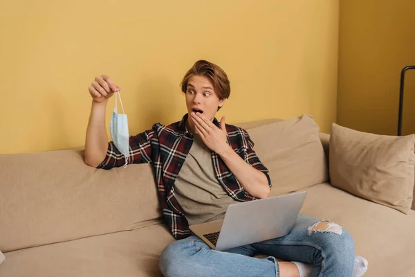 Freelancer sorprendido sosteniendo máscara médica cerca del ordenador portátil en la sala de estar, fin del concepto de cuarentena - foto de stock