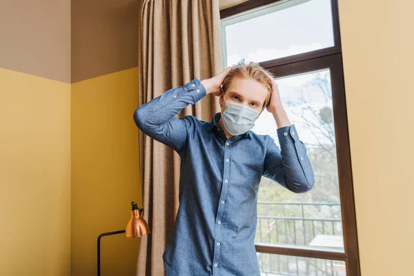 Joven en máscara médica tocando el pelo en casa - foto de stock