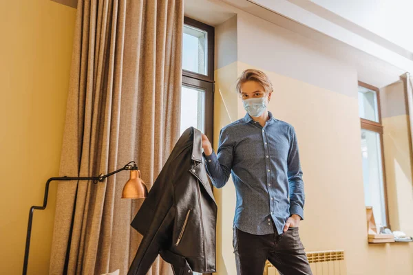 Jovem em máscara médica segurando jaqueta e de pé com a mão no bolso em casa, fim do conceito de quarentena — Fotografia de Stock