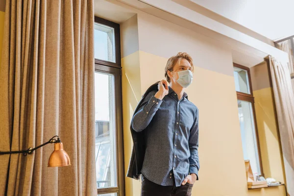 Homem em máscara médica segurando jaqueta e de pé com a mão no bolso em casa, fim do conceito de quarentena — Fotografia de Stock