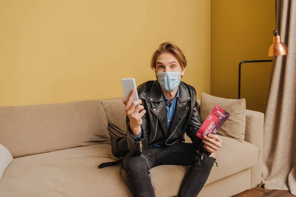 Stylish man in medical mask and jacket holding night party tickets and smartphone, end of quarantine concept — Stock Photo