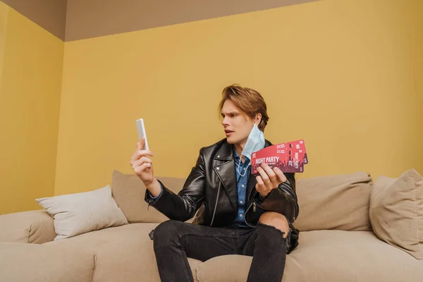 Handsome man in medical mask and jacket holding night party tickets and smartphone, end of quarantine concept — Stock Photo