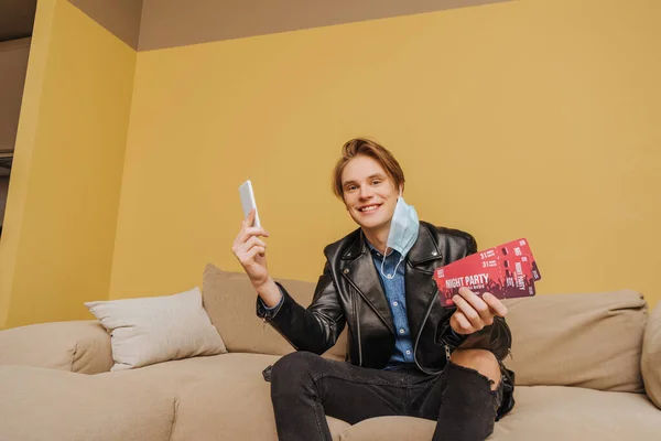 Hombre alegre en máscara médica y chaqueta celebración de entradas de fiesta de noche y teléfono inteligente, fin del concepto de cuarentena - foto de stock