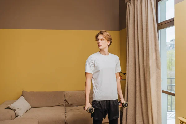 Jovem bonito exercitando com halteres na sala de estar, fim do conceito de quarentena — Fotografia de Stock