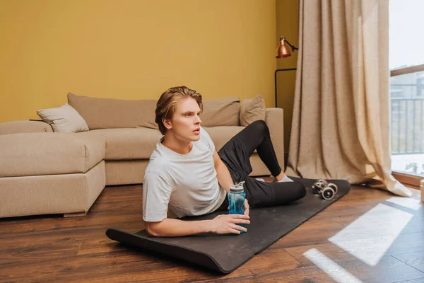 Junger Mann liegt auf Fitnessmatte und hält Sportflasche mit Wasser, Ende der Quarantäne — Stockfoto